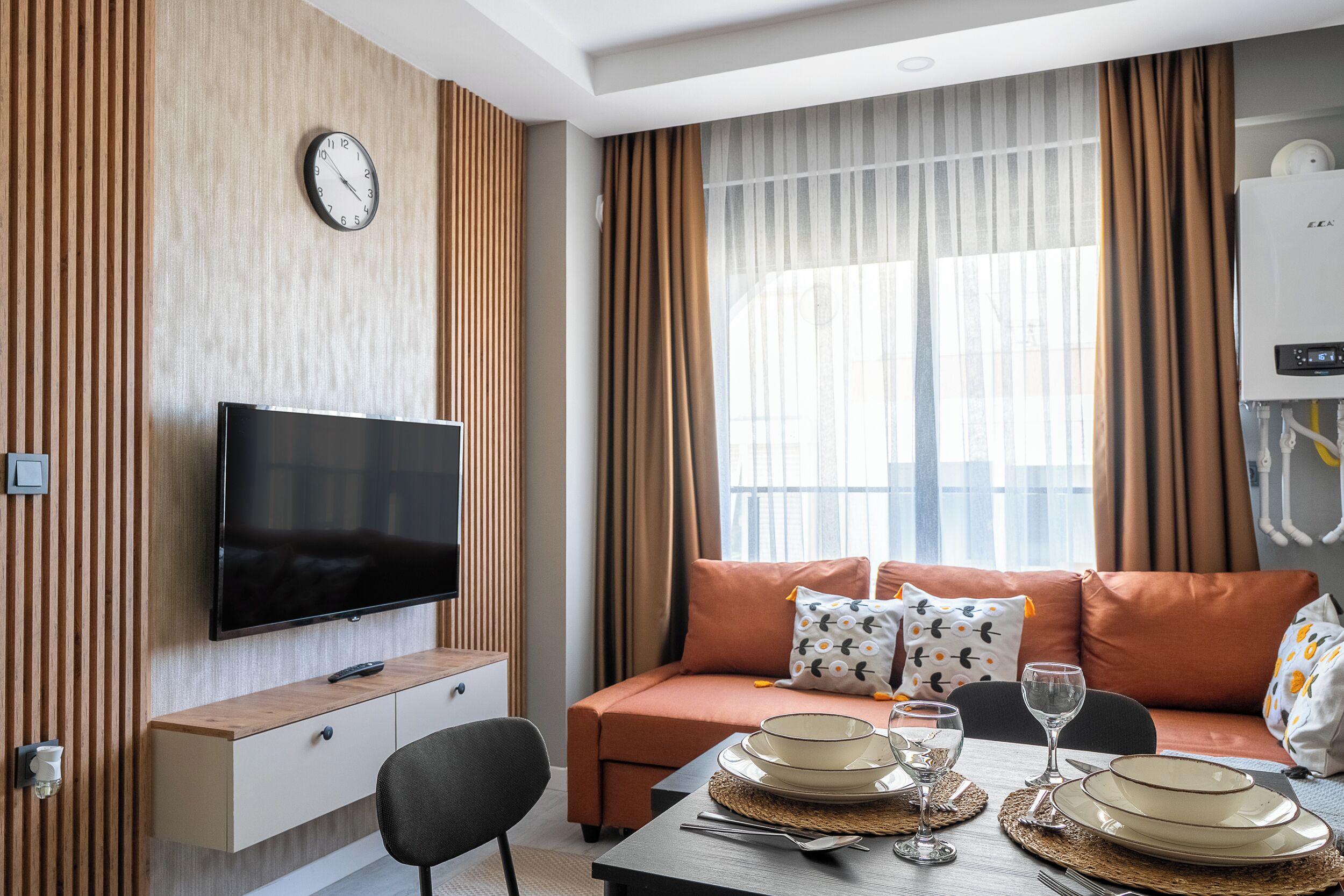 A modern living room with a flat-screen TV, a sofa with patterned cushions, a dining table set with plates and glasses, and a large window with curtains.