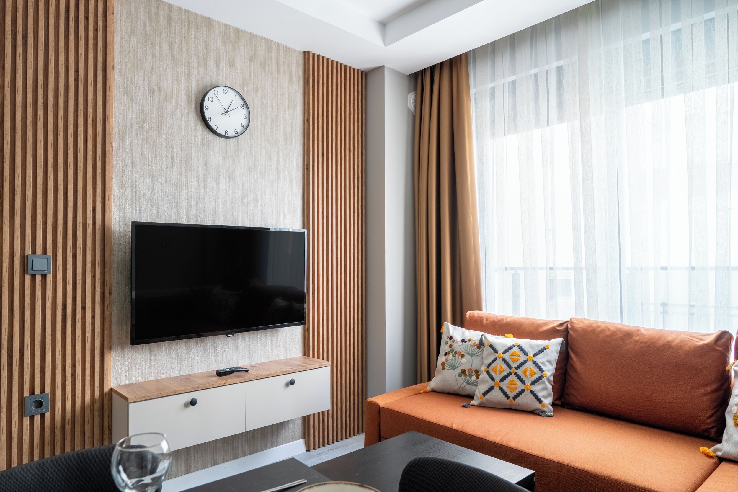 Un soggiorno moderno con TV a schermo piatto, divano con cuscini decorati, parete rivestita in legno e una grande finestra con tende.