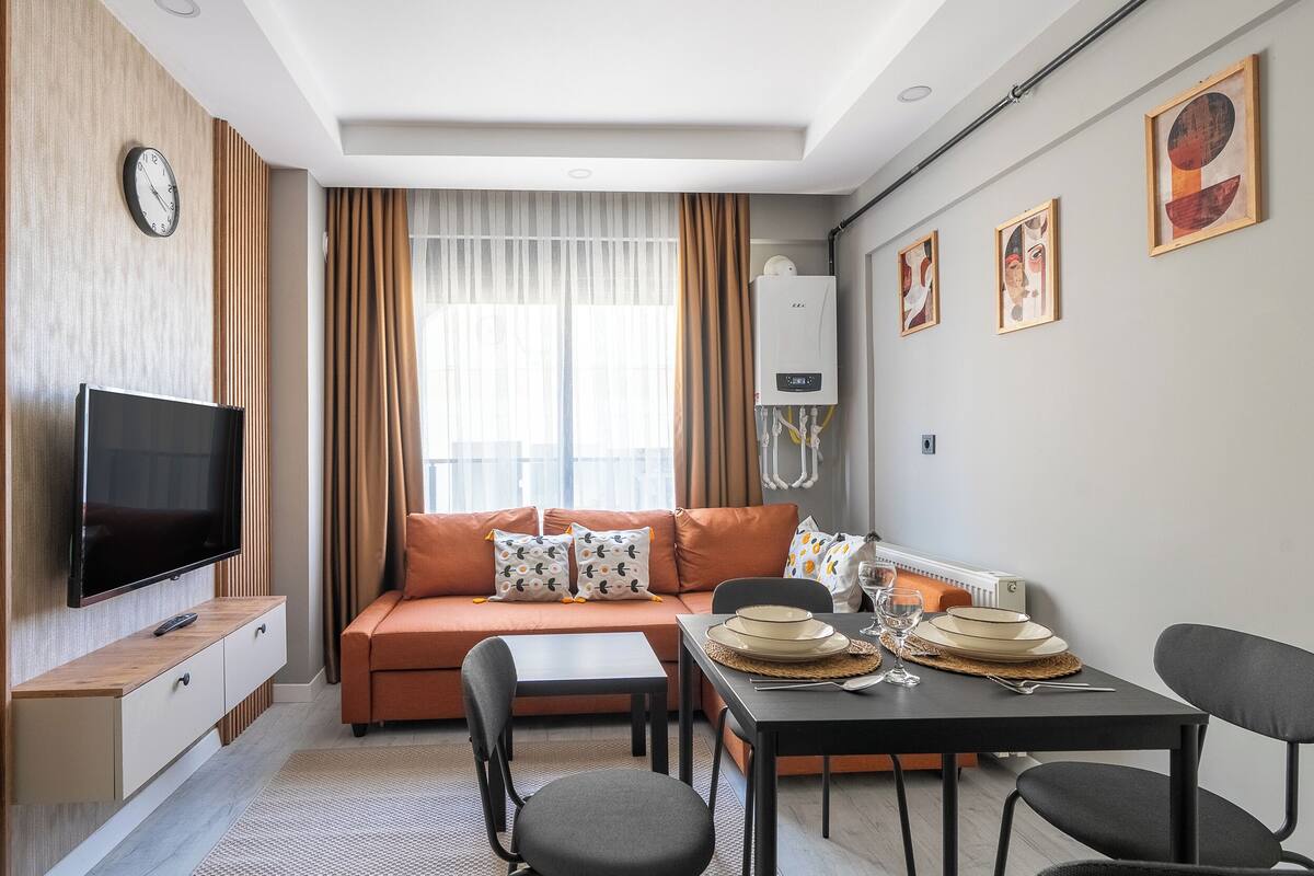 A modern living room with a flat-screen TV, orange sofa, dining table with chairs, and artwork on the wall.