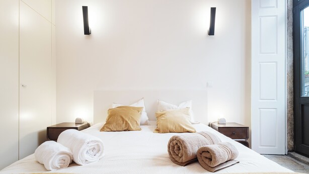 A neatly made bed with rolled towels, two bedside tables, and a door with a view. - Heat of the City: The Cozy Loft (Porto)