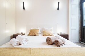 A neatly made bed with rolled towels, two bedside tables, and a door with a view. - Heat of the City: The Cozy Loft (Porto)