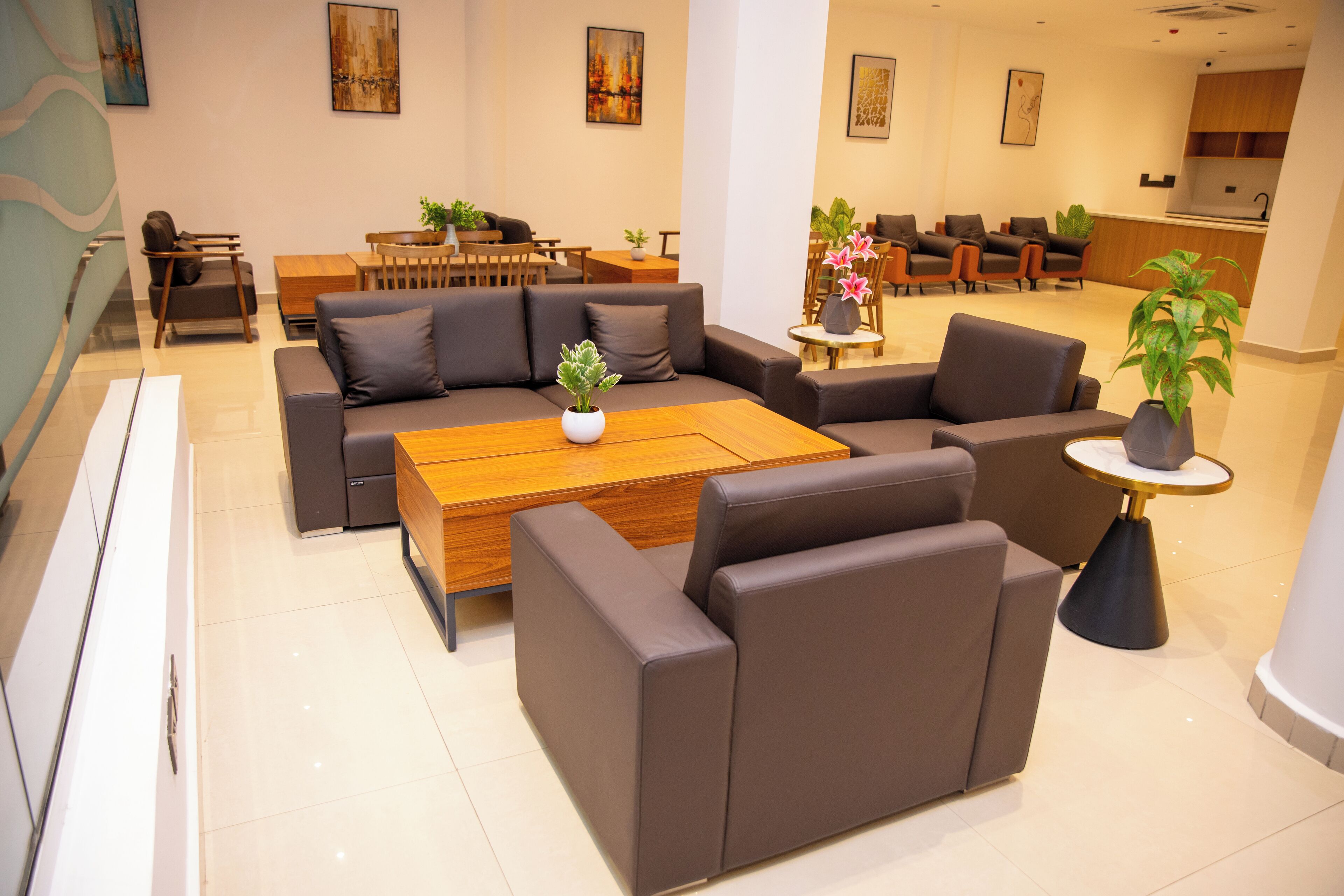 A modern hotel lobby with a sectional sofa, a wooden coffee table, and a small round table with a plant. There are multiple chairs and a reception desk in the background.