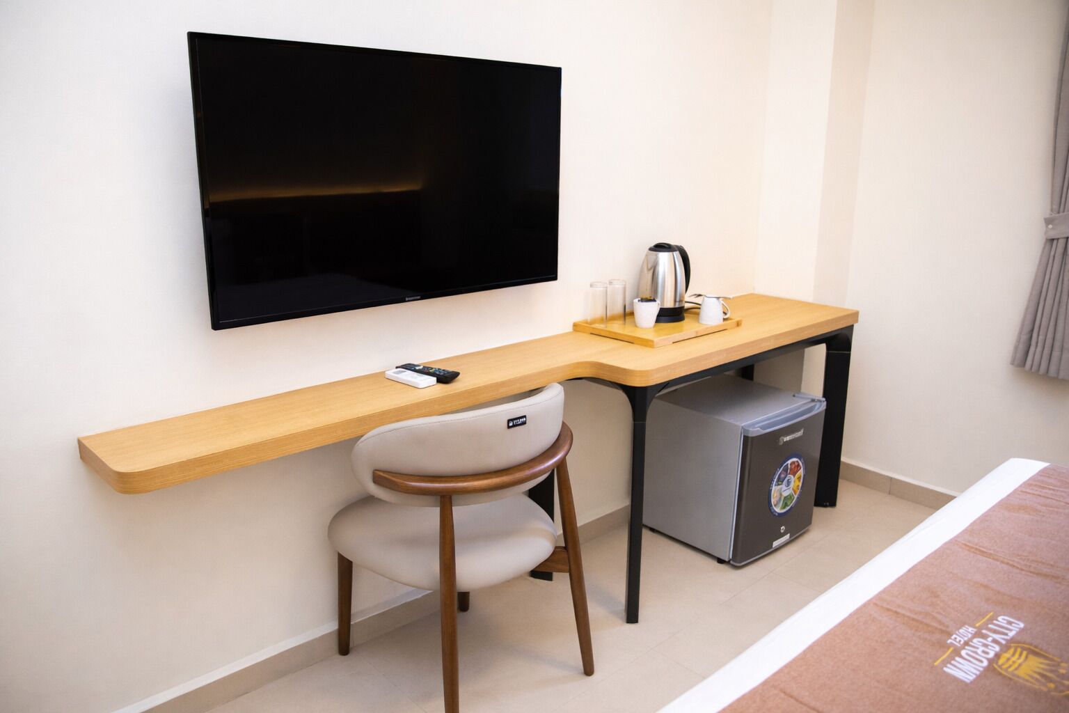 Ett kompakt hotellrum med en platt-TV som är monterad på väggen, ett skrivbord i trä med en stol, ett litet kylskåp och en kaffebryggare.