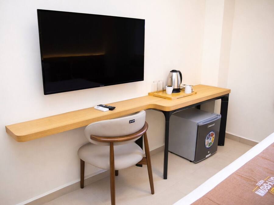 Ett kompakt hotellrum med en platt-TV som är monterad på väggen, ett skrivbord i trä med en stol, ett litet kylskåp och en kaffebryggare.