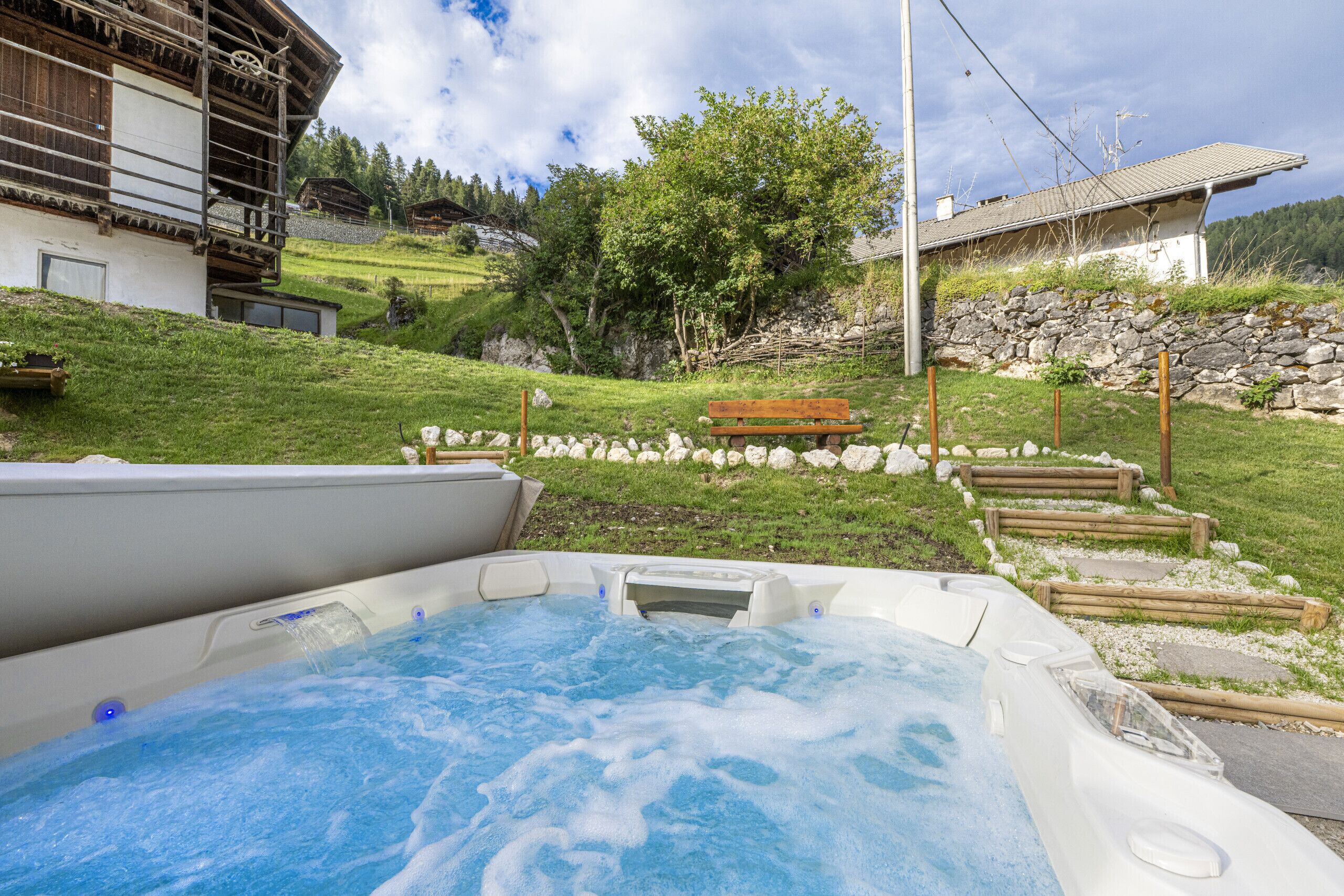 A hot tub in a garden with a wooden bench and stone wall.
