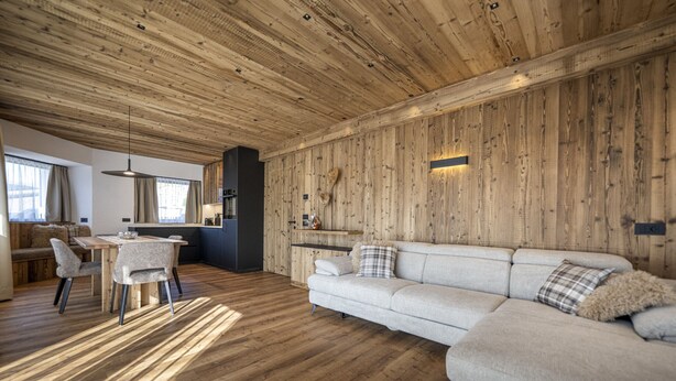 A modern interior with a wooden ceiling, a dining area with a table and chairs, a sofa with cushions, and a kitchen area with a countertop. - 121A - Dosplain Panoramic Apartments | Odles (Santa Cristina Valgardena)