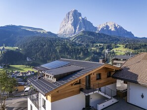 A modern building with a sloped roof, solar panels, and a view of a mountain range. - 121A - Dosplain Panoramic Apartments | Odles (Santa Cristina Valgardena)
