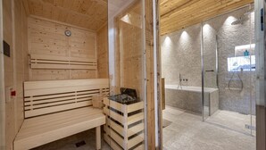 A sauna room with wooden benches, a bench with a stone basin, and a glass shower enclosure. - 121A - Dosplain Panoramic Apartments | Odles (Santa Cristina Valgardena)