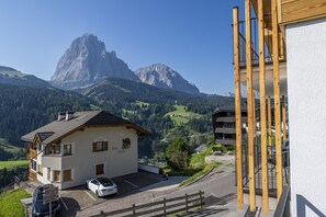 A mountain cabin with a car parked in front, surrounded by lush greenery and a prominent mountain in the background. - 121B - Dosplain Panoramic Apartments | Meisules (Santa Cristina Valgardena)
