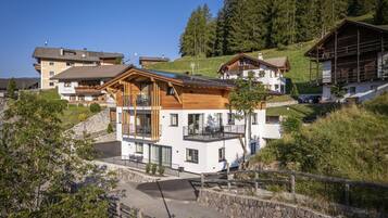 A modern two-story house with a balcony, surrounded by lush greenery and a stone wall.