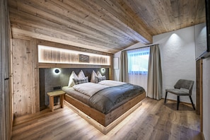 Ein Schlafzimmer mit Holzbalkendecke, einem Bett mit vielen Kissen, einem Nachttisch und einem Sessel.