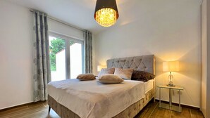 Ett sovrum med en stor säng, en kristallkrona, ett nattduksbord med en lampa och utsikt över naturen genom ett fönster.