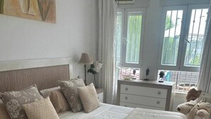 A bedroom with a large bed, a dresser, a lamp, and a window with a view of greenery. - Sun-Drenched Garden Apartment, Castillo Duquesa (Manilva)