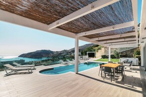 Ein moderner Poolbereich im Freien mit Liegestühlen, einem Holztisch und Blick aufs Meer.