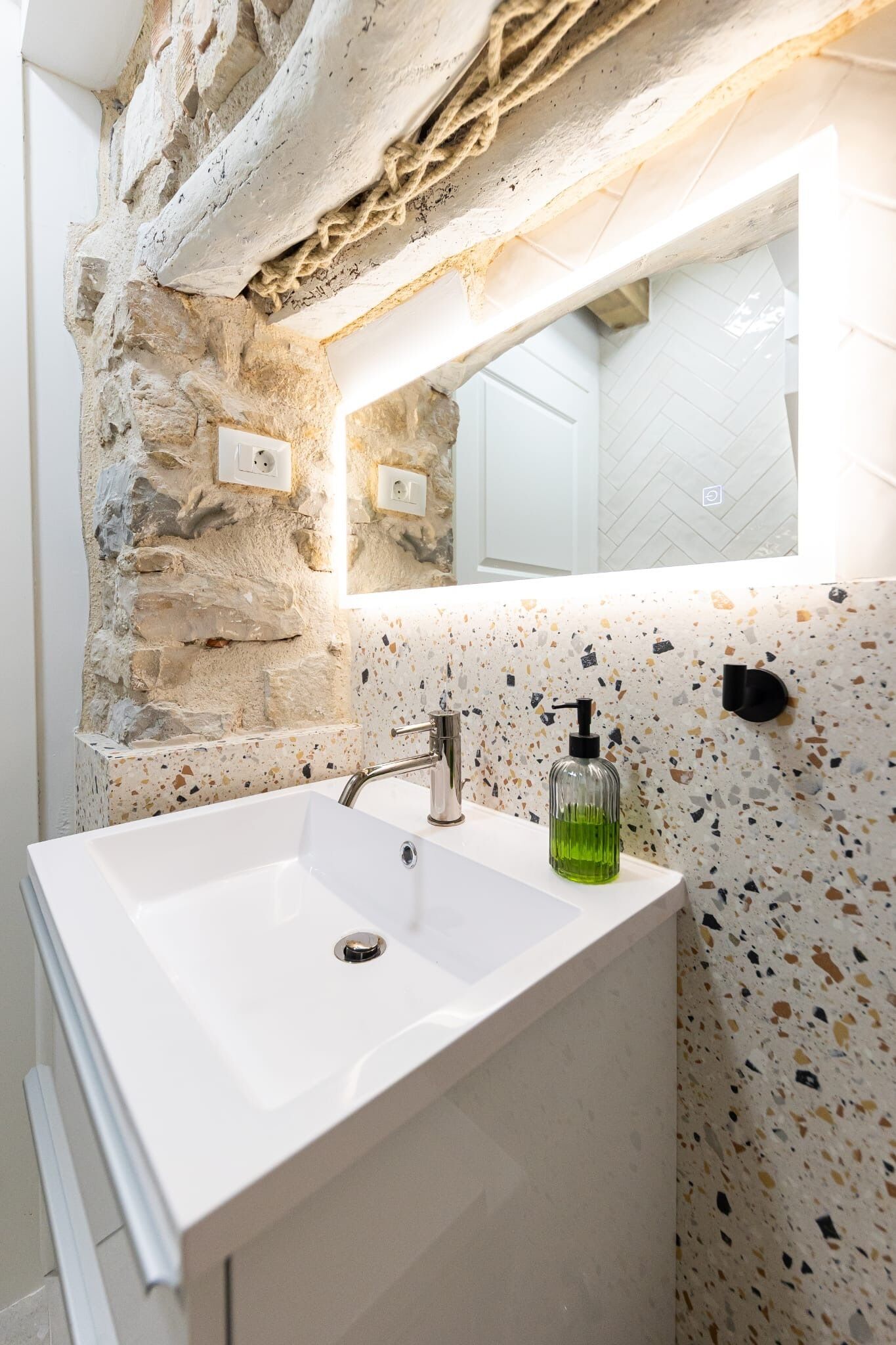 Casa de banho com lavatório branco, parede de pedra e painel de azulejos com manchas.