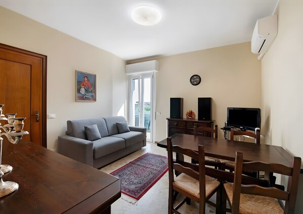 A living room with a sofa, dining table, chairs, a TV, speakers, and a clock. - Apartment 'Solarolo Eleonora' with Balcony and Wi-Fi (Solarolo)