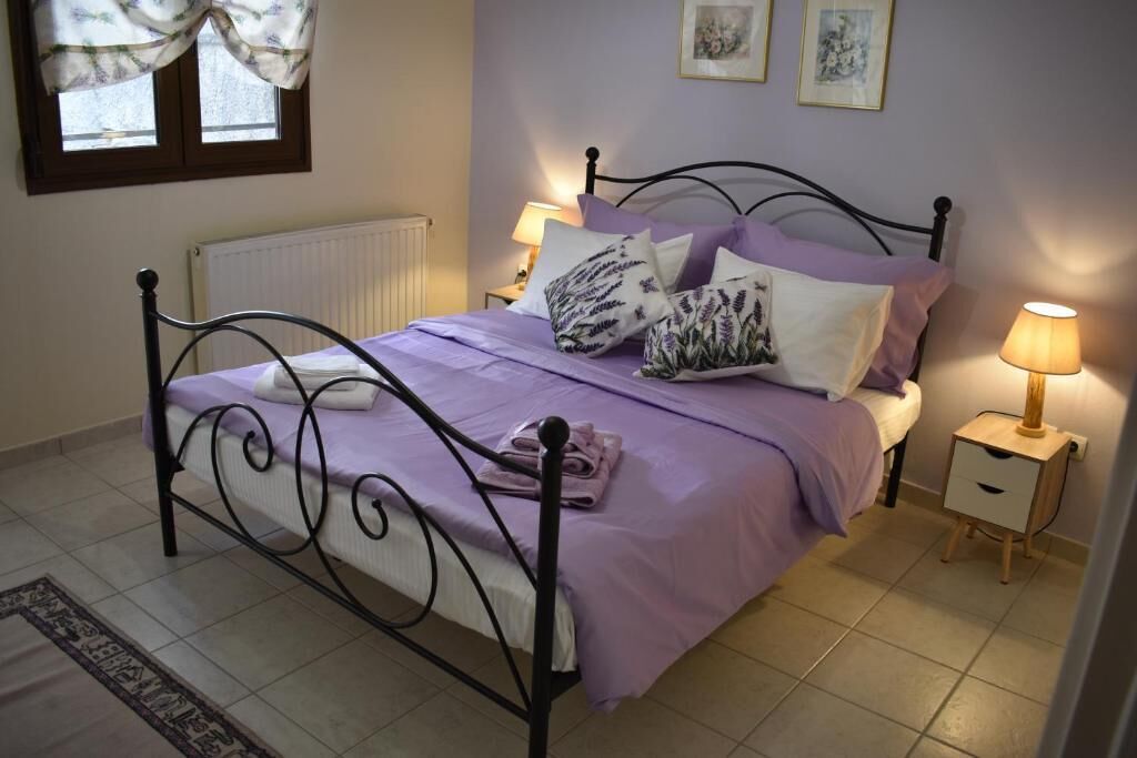 Una cama bien hecha con una colcha morada y almohadas a juego, una mesita de noche con lámpara y una ventana con cortinas.