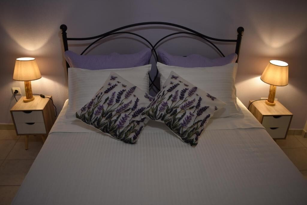 Una cama con estructura metálica negra, ropa de cama blanca y almohadas estampadas con motivos de lavanda. Hay dos mesitas de noche con lámparas a cada lado de la cama.