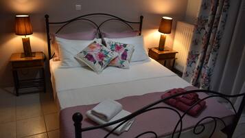 Een netjes opgemaakt bed met bloemenkussens, twee bedlampen, een roze deken en een opgevouwen handdoek op het voeteneinde.