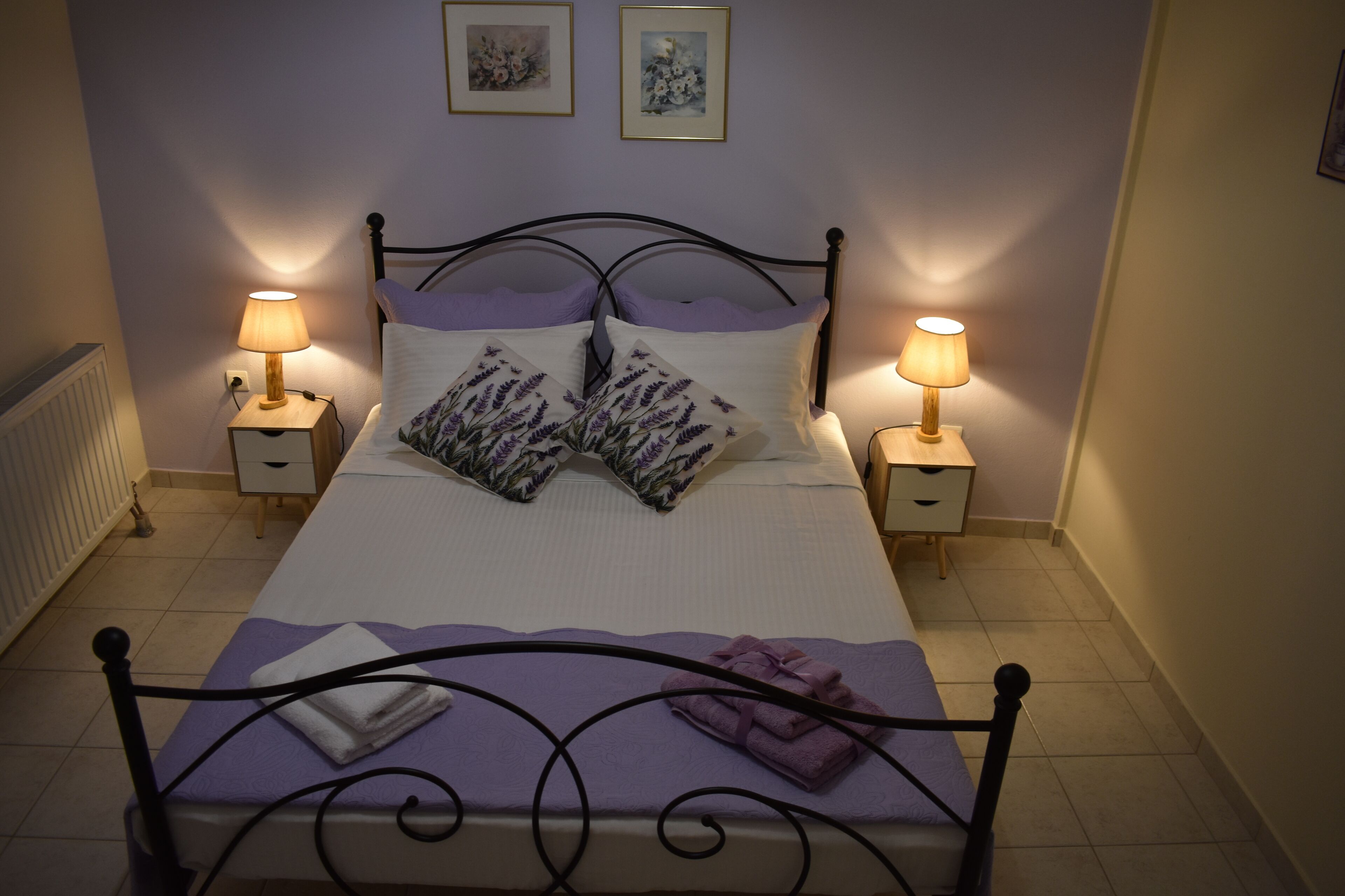 Ein ordentlich gemachtes Bett mit violettem Überwurf, zwei lavendelfarbenen Kissen und einem auf dem Kopfteil gefalteten Handtuch. Zwei Nachttische mit Lampen stehen bereit, und der Raum hat ein schlichtes, sauberes Design.