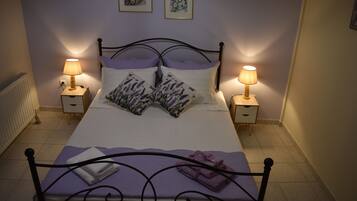 Ein ordentlich gemachtes Bett mit violettem Überwurf, zwei lavendelfarbenen Kissen und einem auf dem Kopfteil gefalteten Handtuch. Zwei Nachttische mit Lampen stehen bereit, und der Raum hat ein schlichtes, sauberes Design.