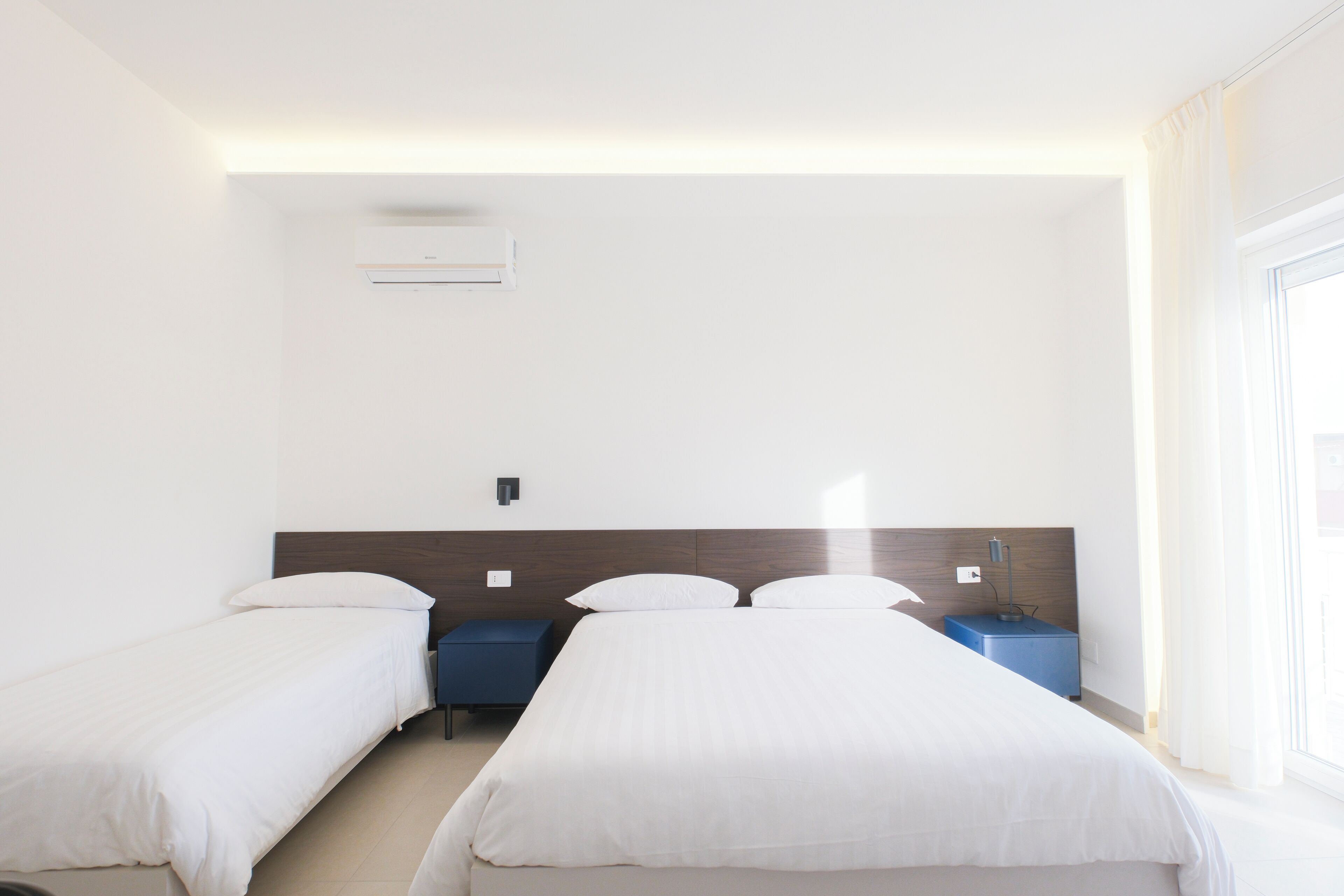Quarto de hotel com duas camas de solteiro, uma mesa de cabeceira, um ar condicionado fixado na parede e uma janela com cortinas.