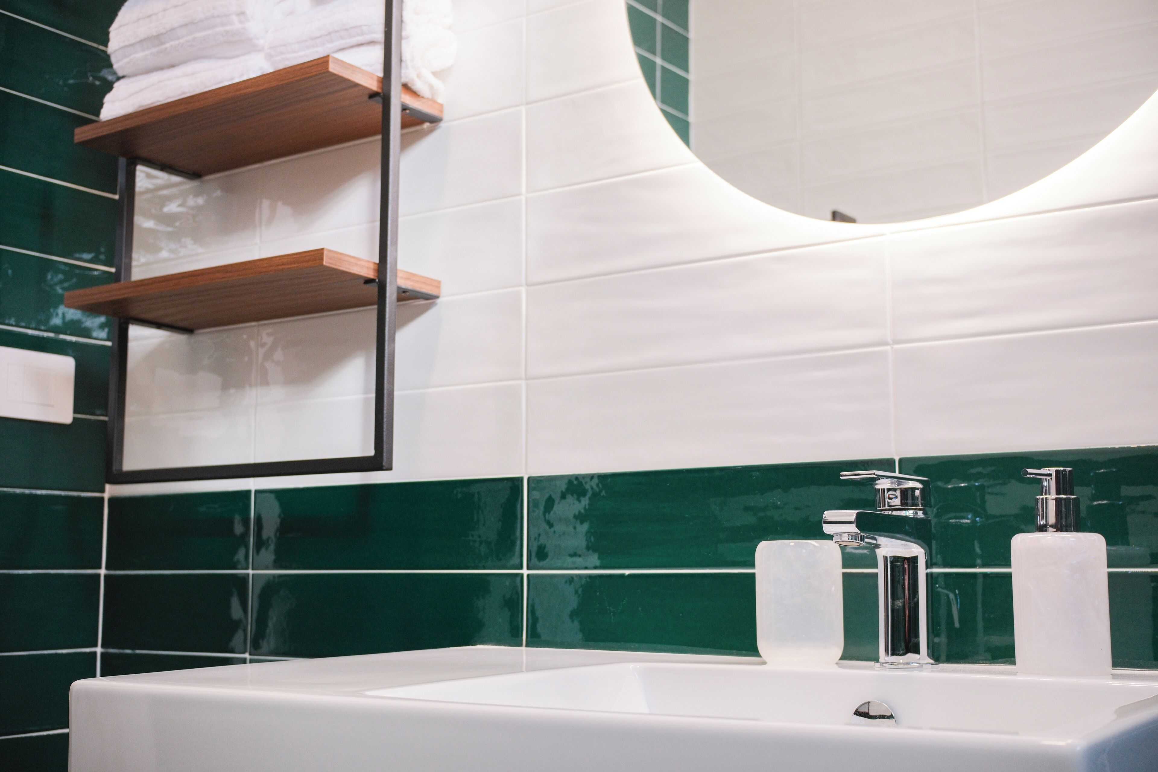 Ein Badezimmer mit einem weißen Waschbecken, zwei Wasserhähnen und einem Spiegel darüber. Auf der linken Seite befinden sich Holzregale mit Handtüchern.