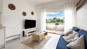 Una sala de estar moderna con un televisor de pantalla plana, una mesa de centro de madera, un sofá con cojines y un amplio ventanal corredero que da a un balcón al aire libre.