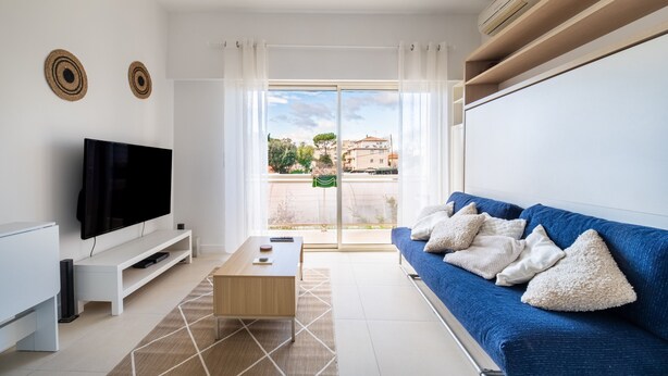 Una sala de estar moderna con un televisor de pantalla plana, un sofá azul con cojines, una mesa de centro de madera y vistas al exterior a través de una puerta de vidrio.