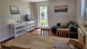 En stue med et træbord, en sofa, et fjernsyn på et hvidt skab og et vindue med udsigt til grønne omgivelser.