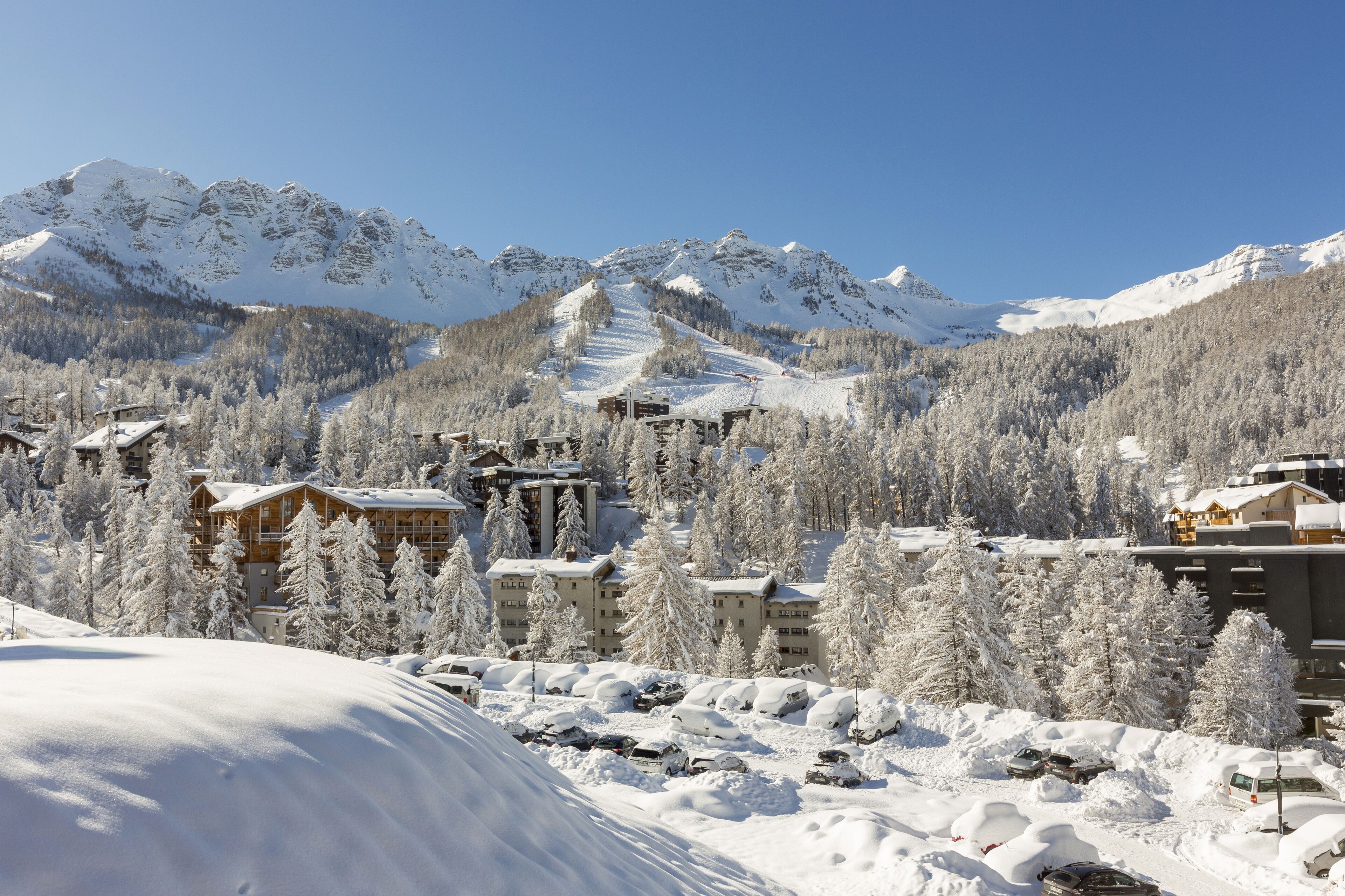 Une station de montagne enneigée, composée de plusieurs bâtiments, dont des chalets et un télésiège, et entourée d’arbres et de montagnes couverts de neige.