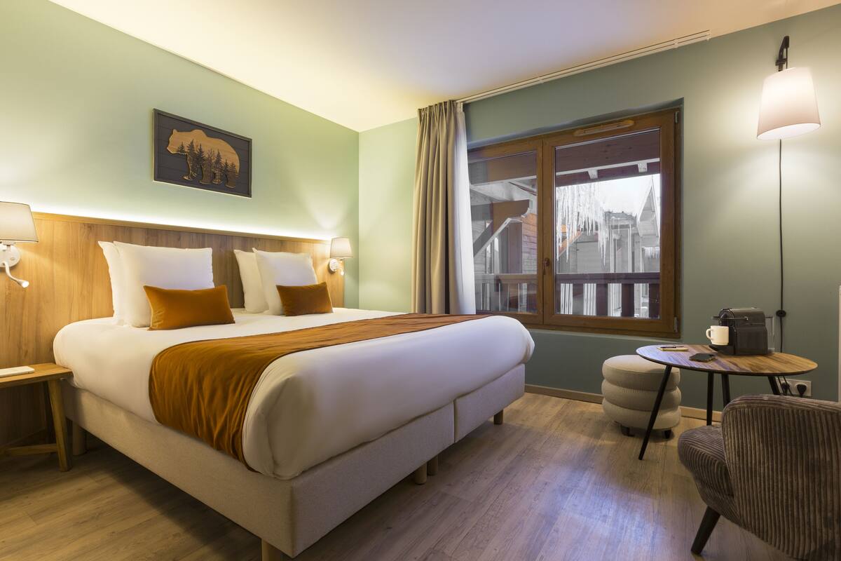 A hotel room with a large bed, a small round table, a chair, a window with a view of buildings, and a lamp.