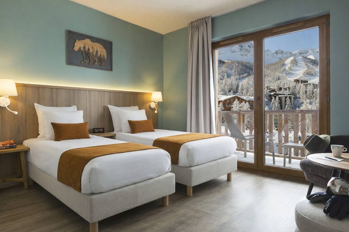A hotel room with two beds, a balcony with a view of snow-covered mountains, and a small table with a chair.