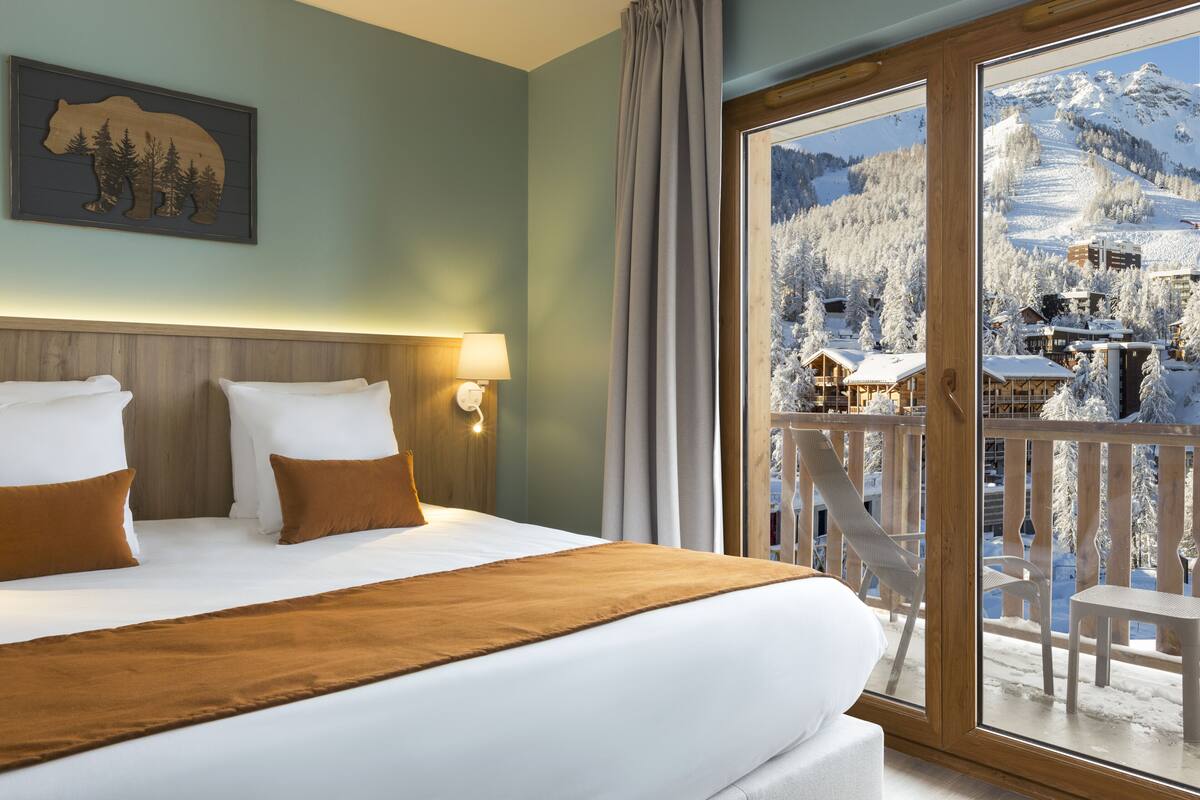 A hotel room with a large bed, a wooden headboard, a mounted bear silhouette, a window with a snowy mountain view, and a balcony with chairs.