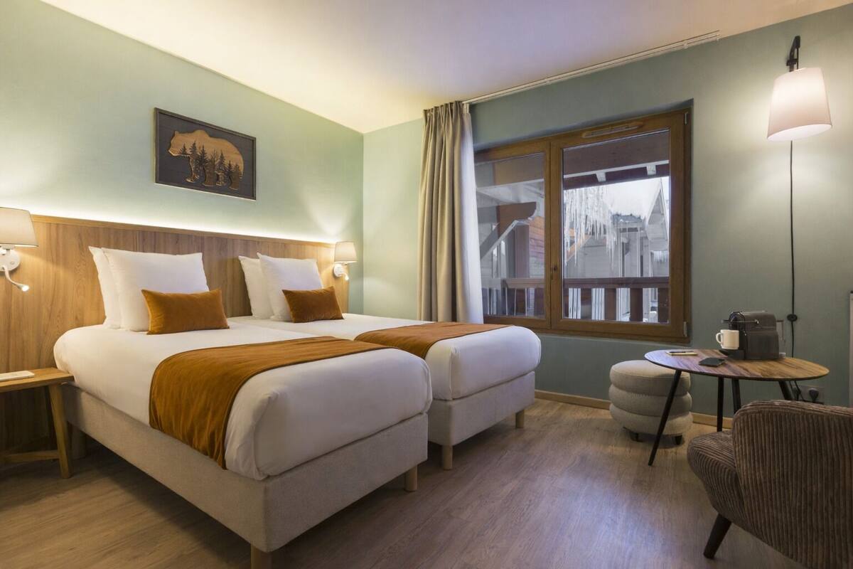 A hotel room with two beds, a small round table, a chair, a window with a view, and a wall-mounted lamp.