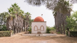 A small, white building with a red dome and two arched windows, surrounded by palm trees and greenery.