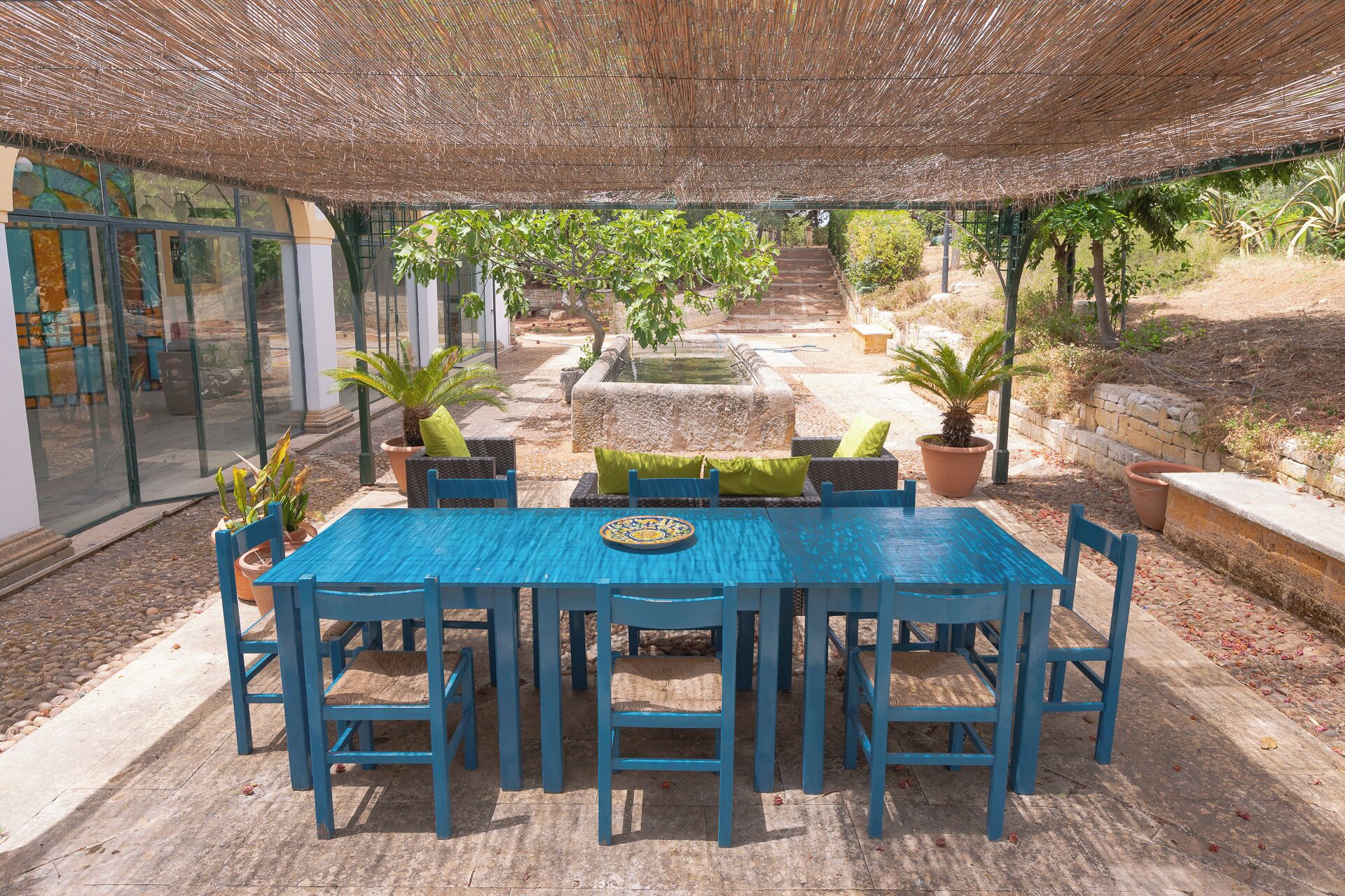 Une terrasse extérieure avec une table et des chaises bleues, entourée de plantes en pot et d’une fontaine en pierre.