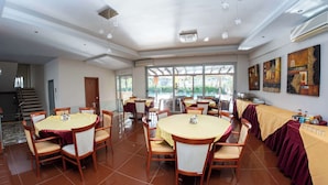 A spacious dining area with round tables, chairs, and a long banquet table covered with a yellow tablecloth. - Hotel Ena - Cozy Mountain View Suite with Pool (Loutra Ypatis)