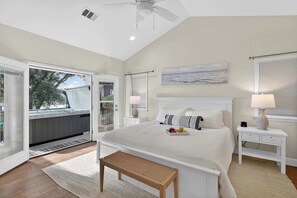 A bedroom with a large bed, a bench, a nightstand, a ceiling fan, and a window with blinds. - Beach House on River Oaks | Concerige Services (Kingsland)