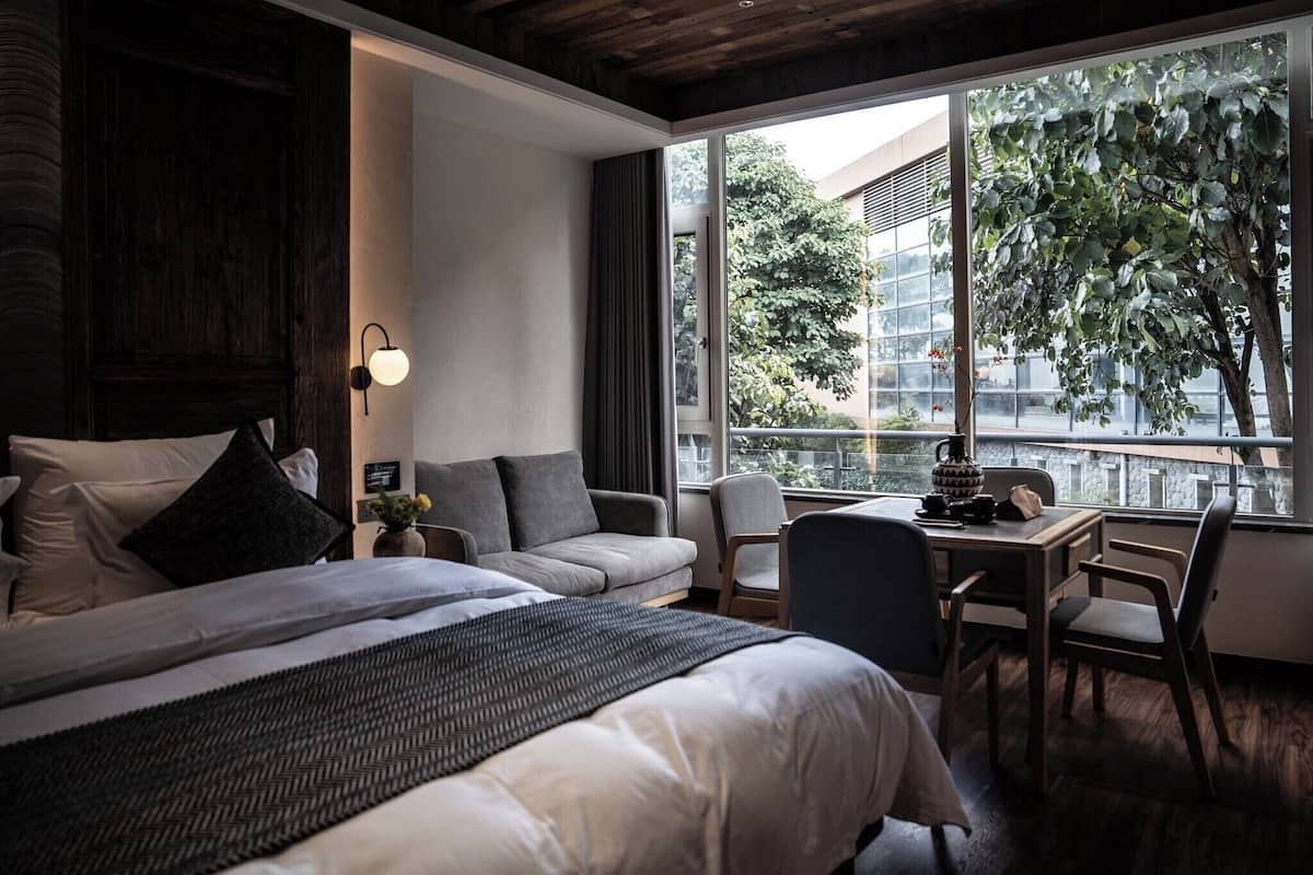 Een moderne hotelkamer met een groot bed, een slaapbank, een eettafel en uitzicht op een stadsgezicht door grote ramen.