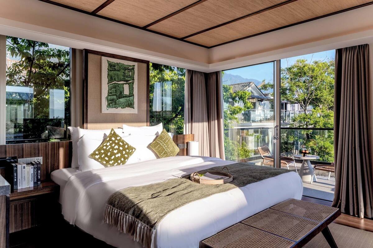 Een moderne hotelkamer met een groot bed, een balkon met uitzicht op groen en traditionele gebouwen, en een houten plafond.