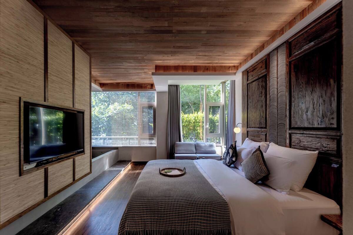 Een moderne slaapkamer met een groot bed, een flatscreen-tv en uitzicht op groen via grote ramen.