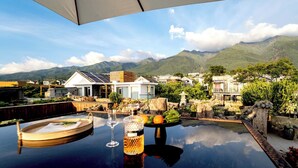 Ein Tisch am Pool mit einem Weinglas, einer Flasche und Obst, mit Blick auf eine Berglandschaft mit modernen Häusern.