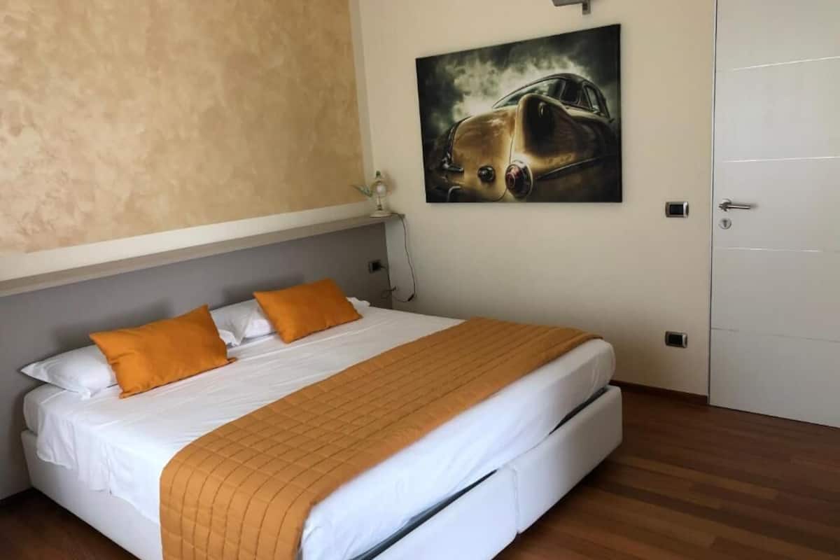 Ein ordentlich bezogenes Bett mit weißer Bettwäsche und einer orangefarbenen Decke, ein Gemälde eines Autos an der Wand und eine Tür rechts.
