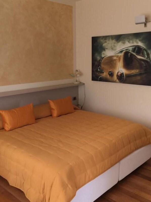 A bedroom with a large bed, orange bedding, a wall-mounted lamp, and a painting of a car. - Giardino Segreto Apartments (Peschiera del Garda)