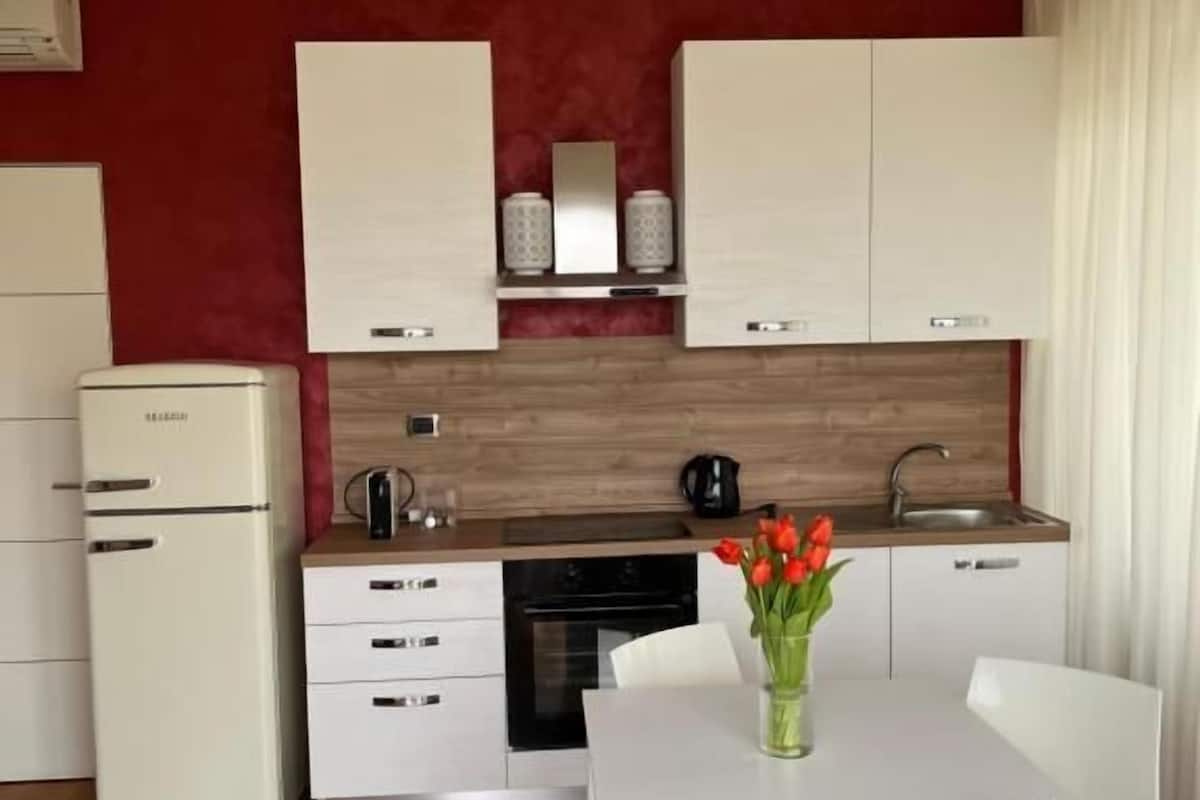 Eine kleine Küche mit weißen Schränken, einem Einbauofen und einer Kaffeemaschine. Ein Esstisch mit Stühlen und eine Vase mit roten Blumen sind ebenfalls vorhanden.