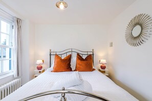 A neatly made bed with white bedding and two orange pillows, flanked by bedside tables with lamps, in a room with a white wall and a decorative mirror. - Candy Cottage | 3-Bed, Walk to Lake & Village (Windermere)