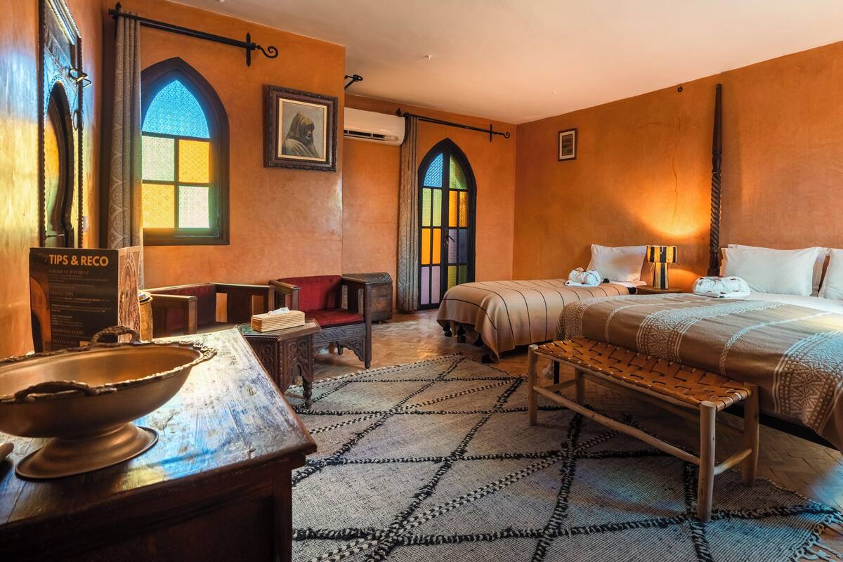 A traditional room with a bed, a wooden bench, a table with a bowl, and two windows with stained glass.