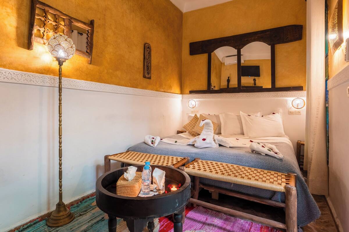 A room with a bed, a small table with a candle, a decorative lamp, and a wall-mounted mirror.