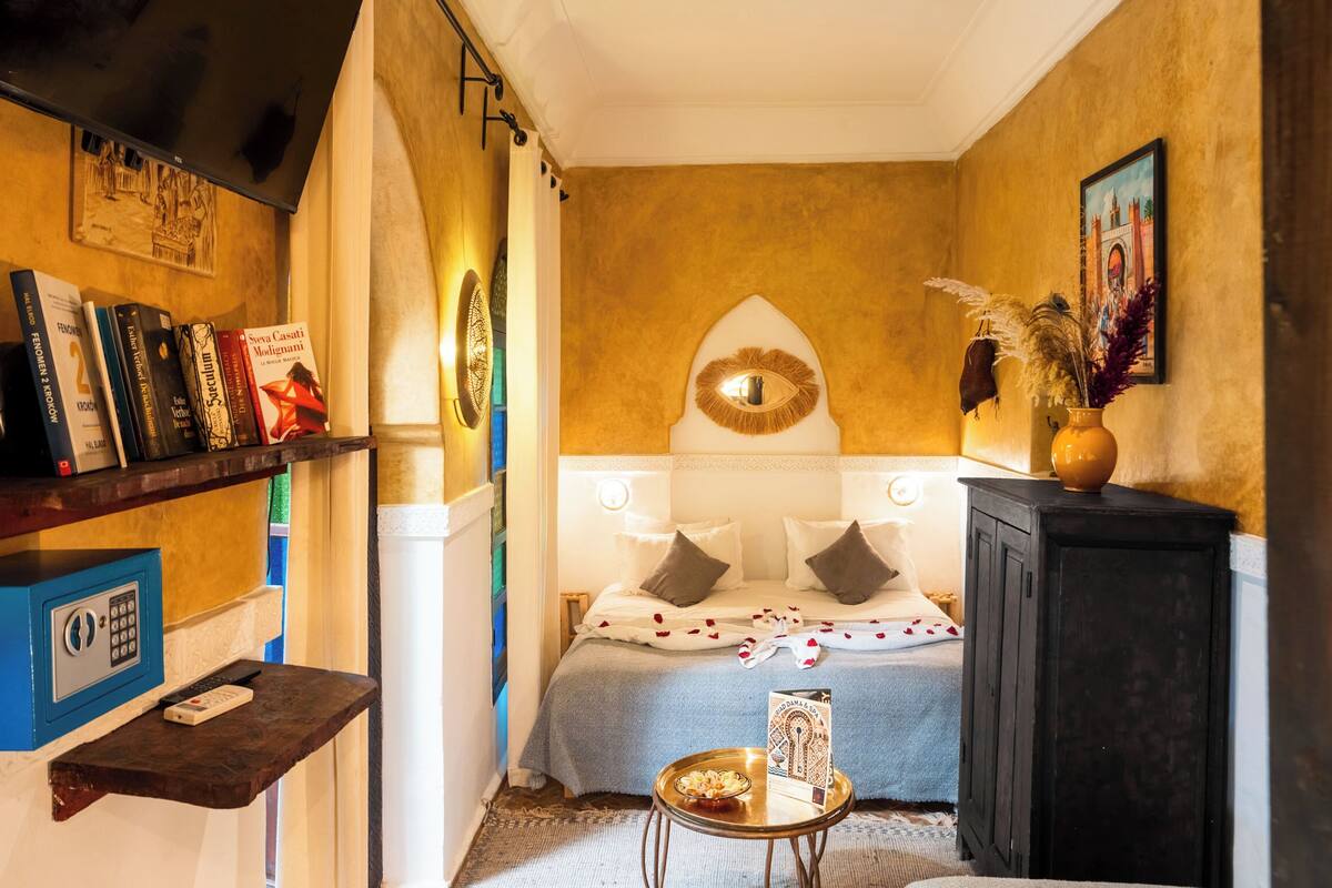 A room with a bed, a small table with a book, a shelf with books, and a wall with a painting.